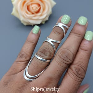 May include: A silver finger ring set. The ring set includes a ring that covers the entire finger, with a curved design and two rings that wrap around the finger. The rings are on a hand with light green nail polish. The text "ShipraJewelry" is at the bottom.