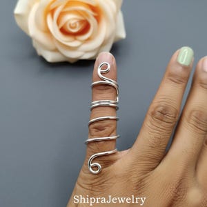 May include: A silver-colored wire ring designed to wrap around the length of a finger. The ring features a continuous spiral pattern with coiled ends at the top and bottom, creating a layered look that extends along the finger joint.