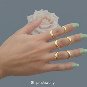 May include: A hand shows four gold rings, each encircling a finger. The rings have a smooth, polished finish. The background features a soft, blurred rose and a muted blue-grey backdrop. The nails are painted a light green colour. The text "ShipraJewelry" is at the bottom.