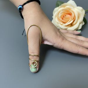 May include: A gold-colored finger ring with a unique design. The ring features a long, curved piece that extends over the top of the finger, with a spiral and two bands around the base. A black bracelet with a blue eye charm is also visible.