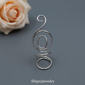 May include: A silver-colored, abstract ring with a spiral design. The ring features a series of loops and curves, with an oval shape in the center. The ring is set against a gray background, with a rose in the upper left corner.