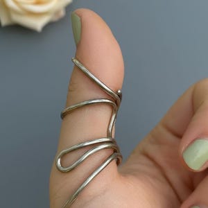 Może przedstawiać: Srebrzysty metalowy pierścień na palec typu szyna z ciągłym drutem, który owija się wokół kciuka w serii pętli. Biżuteria ma gładkie, polerowane wykończenie i jest umieszczona w sposób wspierający strukturę stawu kciuka.