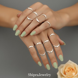 May include: Several silver rings, featuring curved and band designs, adorn two hands with pale green nail polish. The rings are the primary focus, highlighting a minimalist jewellery style. The brand name "ShipraJewelry" is visible at the bottom of the image.