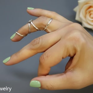 May include: A hand wearing multiple silver rings. The rings include a simple band, a wrap-around design, and a ring that connects to the knuckle. The nails are painted a light green color. A rose is in the background.