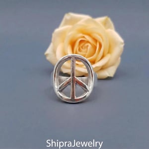 May include: A silver ring displaying the peace symbol, set against a soft grey backdrop with a yellow rose in the background. The ring's design is a bold, circular shape with a vertical and two diagonal lines. The shop name "ShipraJewelry" is at the bottom.
