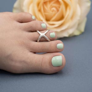 May include: A silver toe ring with an X design is worn on a foot with mint green nail polish. A pale yellow rose is in the background. The text "ShipraJewelry" is at the bottom.