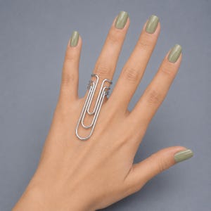 Peut inclure: Une bague argentée au design inspiré d'un trombone, constituée d'une boucle de fil métallique continu qui s'étend verticalement le long du doigt. L'anneau métallique présente une finition polie et une forme allongée rappelant la papeterie.