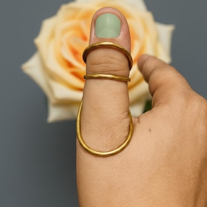May include: Three gold-toned rings worn on a thumb. Two thin, circular bands are positioned near the nail, while a larger, elongated oval-shaped ring encircles the base of the thumb. The metal has a matte, textured finish throughout the design.