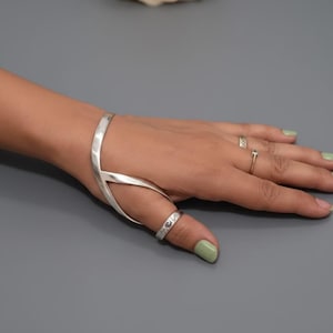 May include: A silver hand bracelet and ring set. The bracelet wraps around the hand and connects to a ring on the thumb. Additional rings are on other fingers. The background is a solid gray with a rose.