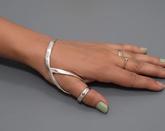 May include: A silver hand bracelet and ring set. The bracelet wraps around the hand and connects to a ring on the thumb. Additional rings are on other fingers. The background is a solid gray with a rose.