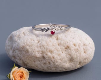 May include: A silver ring featuring a delicate leaf-shaped band and a single small, round, vibrant red gemstone set at the center. The ring is positioned on a textured, light-colored stone, with a small, pale peach-colored rose nearby.