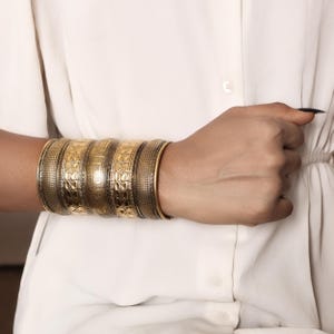 Puede incluir: Un conjunto de brazaletes dorados y bronce adorna una muñeca. Los brazaletes presentan patrones intrincados, incluyendo diseños florales y geométricos. La muñeca se destaca sobre un fondo blanco, resaltando los detalles de la joyería.