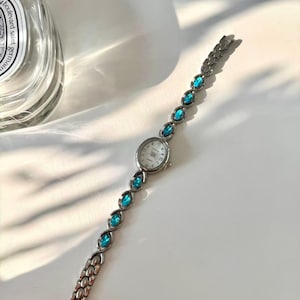 May include: Silver quartz watch with a white face and teal gemstone accents. The watch band features oval teal stones. The watch is approximately 18 cm long.