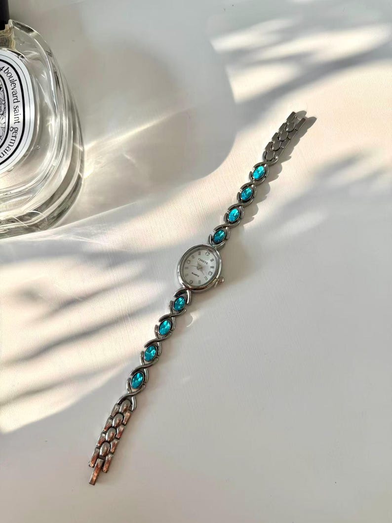 May include: Silver quartz watch with a white face and teal gemstone accents. The watch band features oval teal stones. The watch is approximately 18 cm long.
