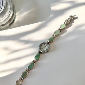 May include: A silver wristwatch featuring a white watch face and a series of oval, pale green stones. The watch strap is made of linked sections, with the green stones set in the middle of each link. The watch has a classic, elegant design.