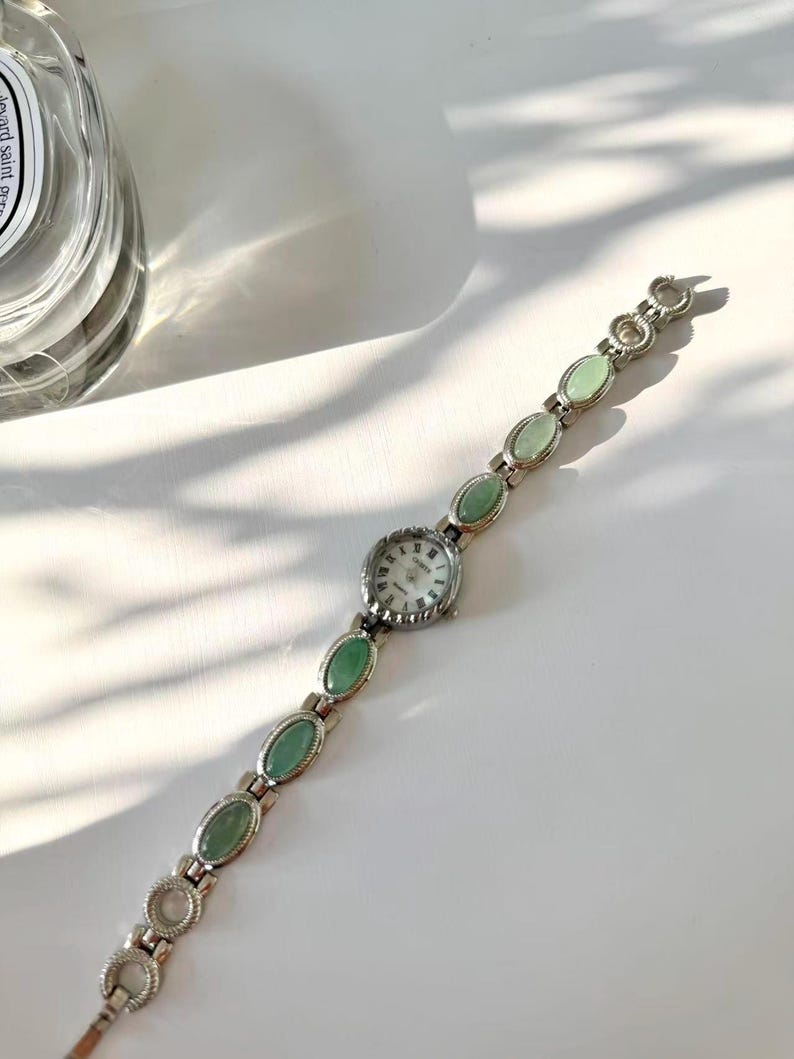May include: A silver wristwatch featuring a white watch face and a series of oval, pale green stones. The watch strap is made of linked sections, with the green stones set in the middle of each link. The watch has a classic, elegant design.