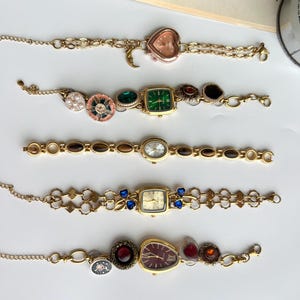 Puede incluir: Cinco pulseras doradas de estilo vintage con esferas de reloj decorativas. Las esferas de los relojes tienen varias formas, incluyendo coraz&oacute;n, cuadrado y ovalado, y presentan diferentes colores y adornos como gemas y perlas. Las pulseras se exhiben sobre una superficie blanca.