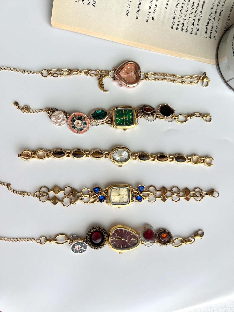 Puede incluir: Cinco pulseras doradas de estilo vintage con esferas de reloj decorativas. Las esferas de los relojes tienen varias formas, incluyendo coraz&oacute;n, cuadrado y ovalado, y presentan diferentes colores y adornos como gemas y perlas. Las pulseras se exhiben sobre una superficie blanca.