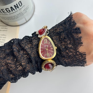 Puede incluir: Un reloj de pulsera dorado con esfera roja y manecillas doradas. El reloj tiene una forma de l&aacute;grima &uacute;nica y se combina con un anillo de piedra roja a juego. El reloj est&aacute; sujeto a una manga de encaje negro.