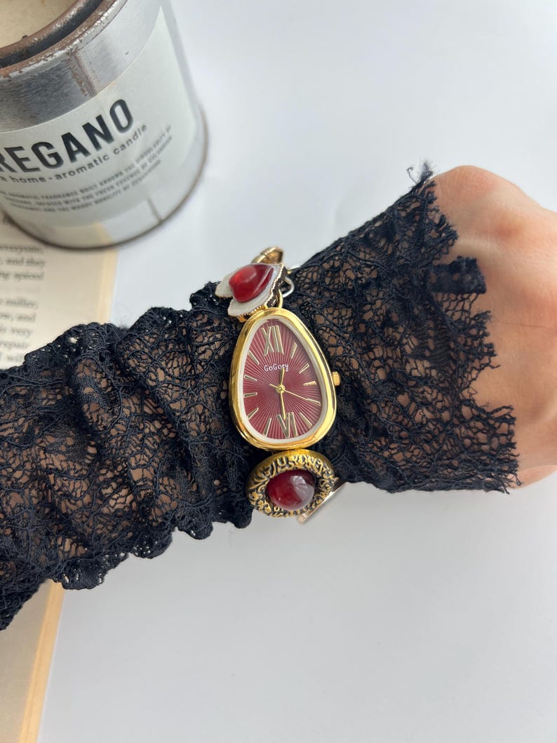 Puede incluir: Un reloj de pulsera dorado con esfera roja y manecillas doradas. El reloj tiene una forma de l&aacute;grima &uacute;nica y se combina con un anillo de piedra roja a juego. El reloj est&aacute; sujeto a una manga de encaje negro.