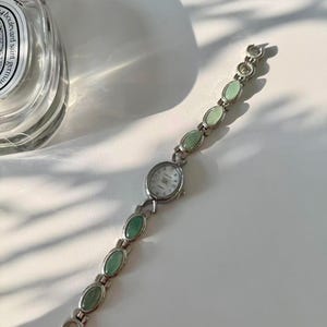 May include: A silver wristwatch featuring a white watch face and a band adorned with oval, pale green stones. The watch has a classic design, suitable for everyday wear.