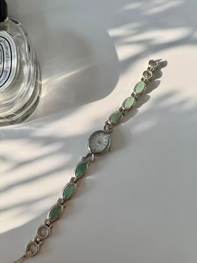 May include: A silver wristwatch featuring a white watch face and a band adorned with oval, pale green stones. The watch has a classic design, suitable for everyday wear.
