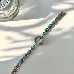 May include: A silver wristwatch with a white face and a decorative bezel. The watch strap features teal-coloured, oval-shaped stones. The watch is displayed on a white surface, highlighting its elegant design.
