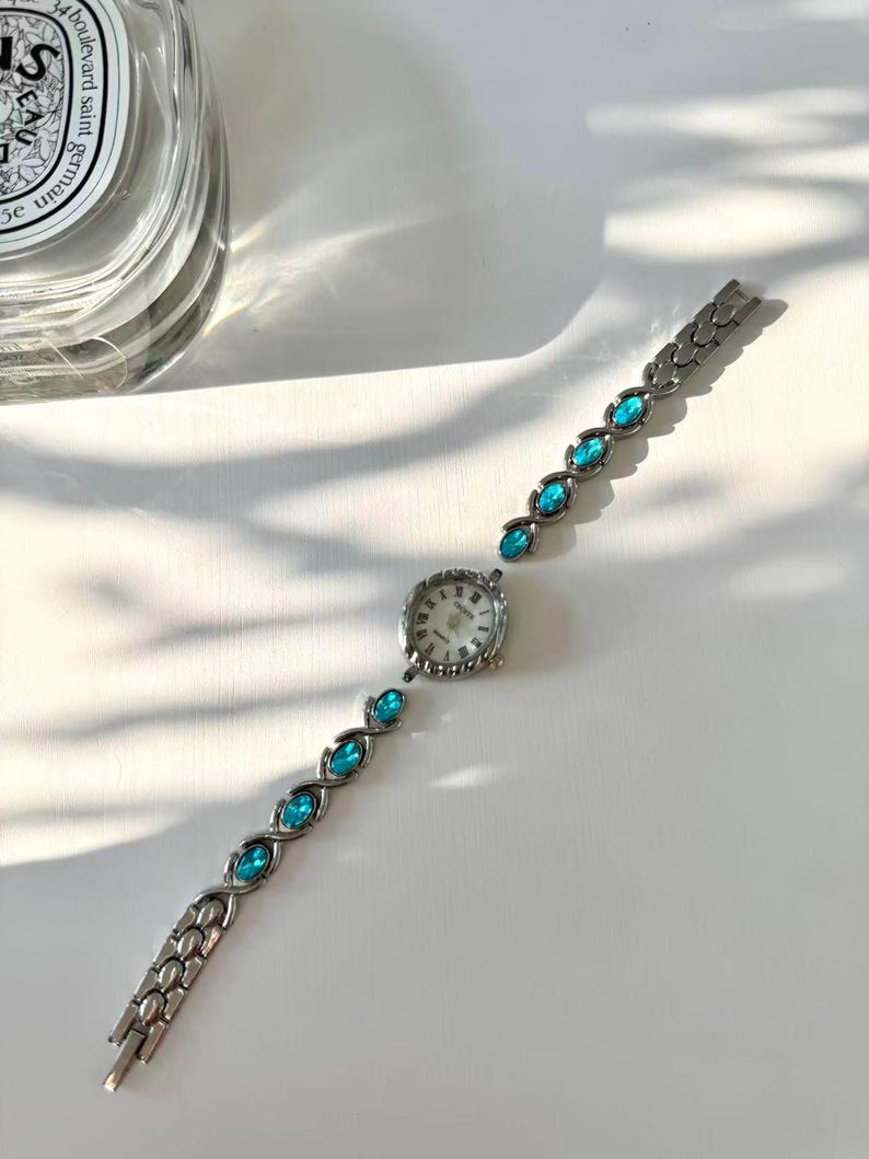 May include: A silver wristwatch with a white face and a decorative bezel. The watch strap features teal-coloured, oval-shaped stones. The watch is displayed on a white surface, highlighting its elegant design.