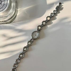 May include: Silver wristwatch with an oval face and a white dial. The watch strap has a series of linked, open circles, each holding a small, round, white pearl. The watch is on a white surface.
