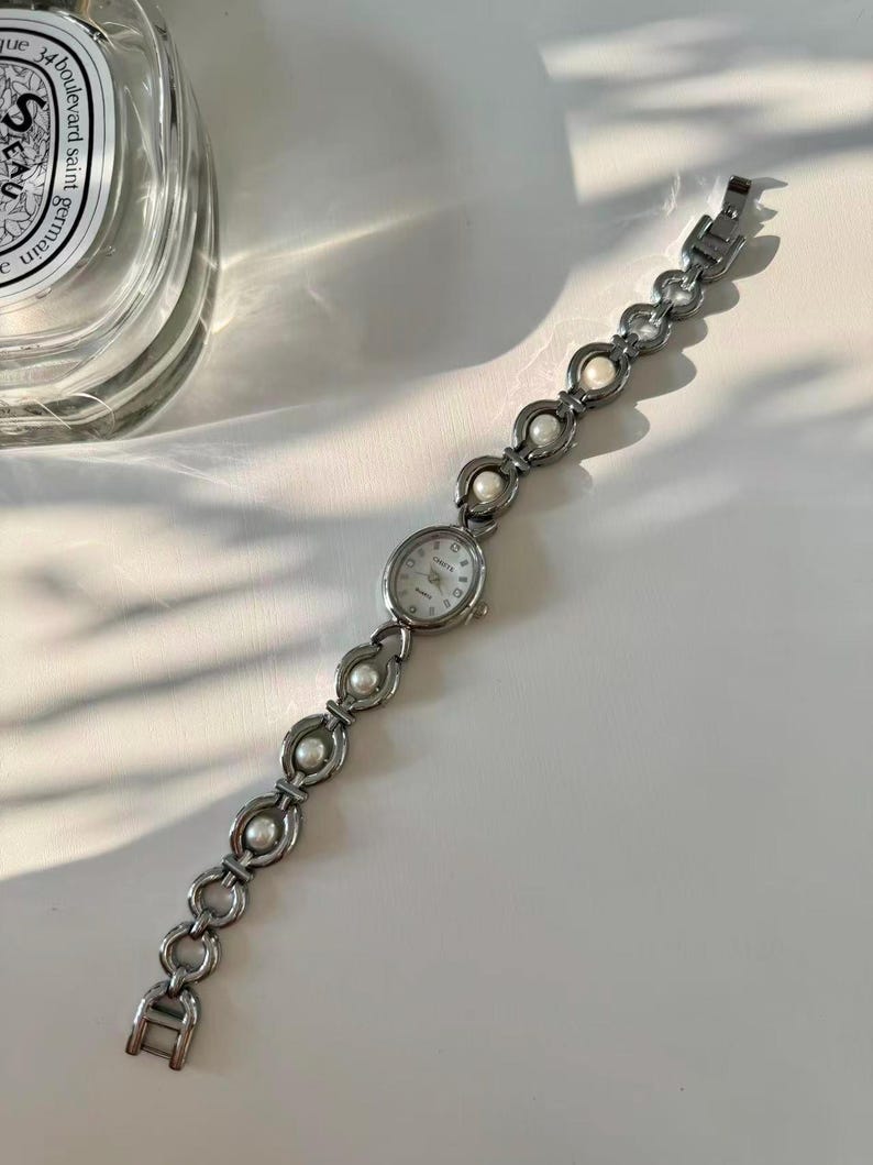 May include: Silver wristwatch with an oval face and a white dial. The watch strap has a series of linked, open circles, each holding a small, round, white pearl. The watch is on a white surface.