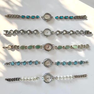 May include: Six vintage wristwatches with silver metal straps. The watch faces are round or octagonal, with details like blue gemstones, faux pearls, and green oval stones. The watches are displayed on a white surface, showcasing their different designs.