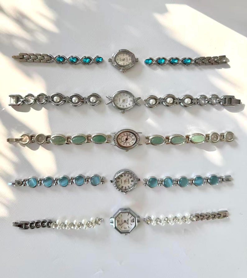 May include: Six vintage wristwatches with silver metal straps. The watch faces are round or octagonal, with details like blue gemstones, faux pearls, and green oval stones. The watches are displayed on a white surface, showcasing their different designs.