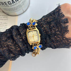 Puede incluir: Un reloj de pulsera dorado con una esfera rectangular y una correa dorada adornada con detalles de joyas azules. La esfera del reloj muestra n&uacute;meros romanos. El reloj se lleva en una manga de encaje negro.