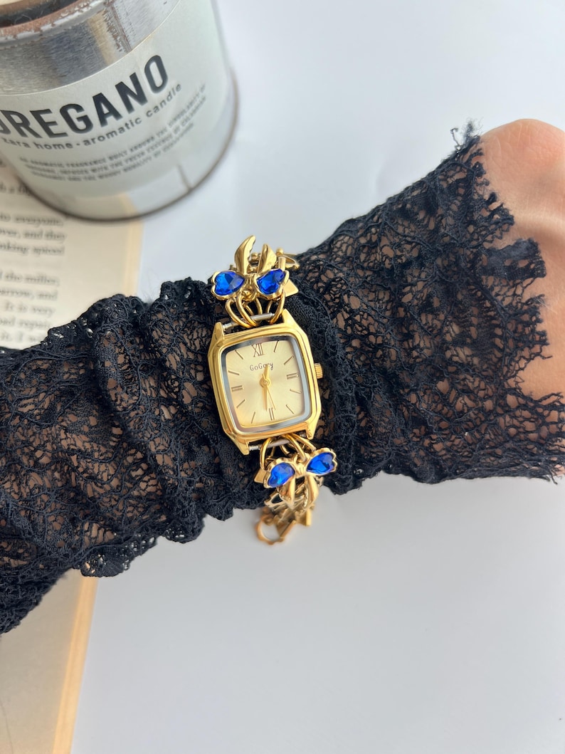 Puede incluir: Un reloj de pulsera dorado con una esfera rectangular y una correa dorada adornada con detalles de joyas azules. La esfera del reloj muestra n&uacute;meros romanos. El reloj se lleva en una manga de encaje negro.