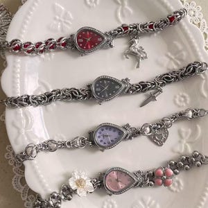 May include: Four teardrop-shaped wristwatches with silver-tone chain link bands. The top watch has a red face and a unicorn charm. The second has a black face and a dagger charm. The third has a light purple face and heart charm. The bottom has a pink face with a flower accent.