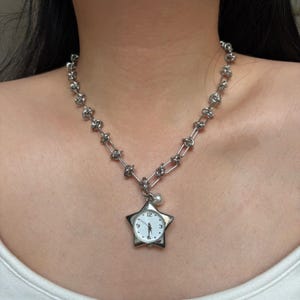 Puede incluir: Collar plateado con un colgante en forma de estrella que contiene un reloj. El reloj tiene una esfera blanca con números y agujas negras. La cadena está hecha de eslabones interconectados. Una pequeña perla cuelga debajo del reloj.