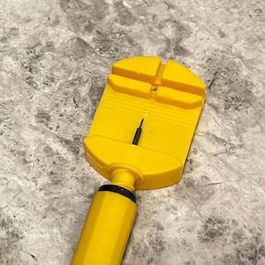 May include: A yellow watch band link remover tool with a black pin. The tool has a rectangular head with four slots and a cylindrical handle. The handle has a black cap. The tool is designed for removing links from watch straps.