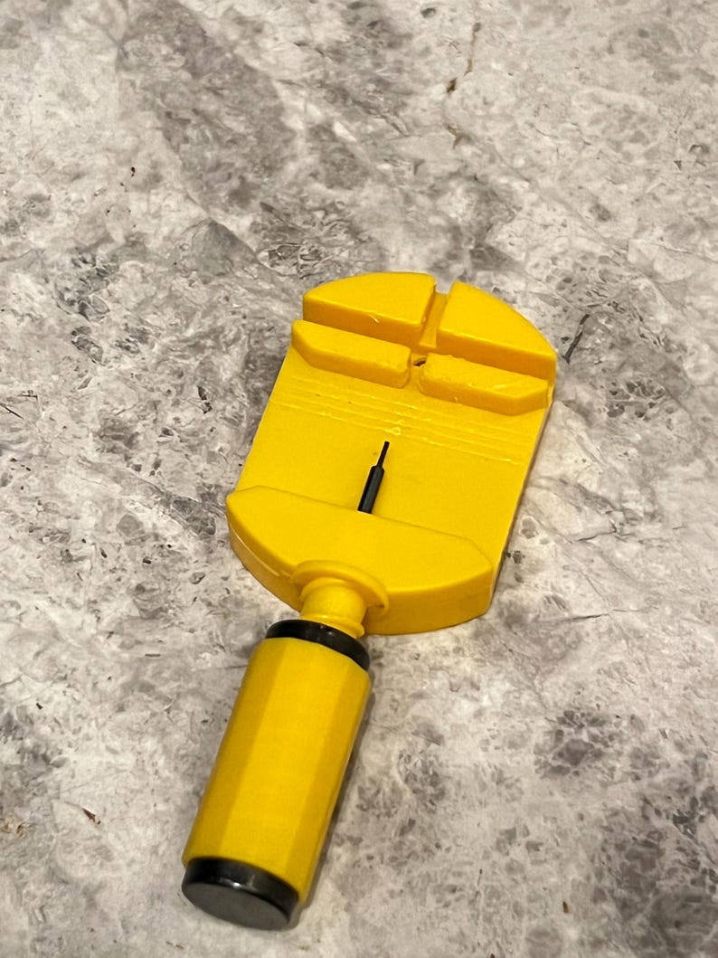 May include: A yellow watch band link remover tool with a black pin. The tool has a rectangular head with four slots and a cylindrical handle. The handle has a black cap. The tool is designed for removing links from watch straps.