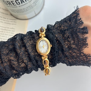 Puede incluir: Un reloj de pulsera dorado con esfera ovalada y esfera blanca. El reloj presenta piedras decorativas marrones y una correa dorada. El reloj se lleva en una manga de encaje negro. Una vela y un libro est&aacute;n en el fondo.