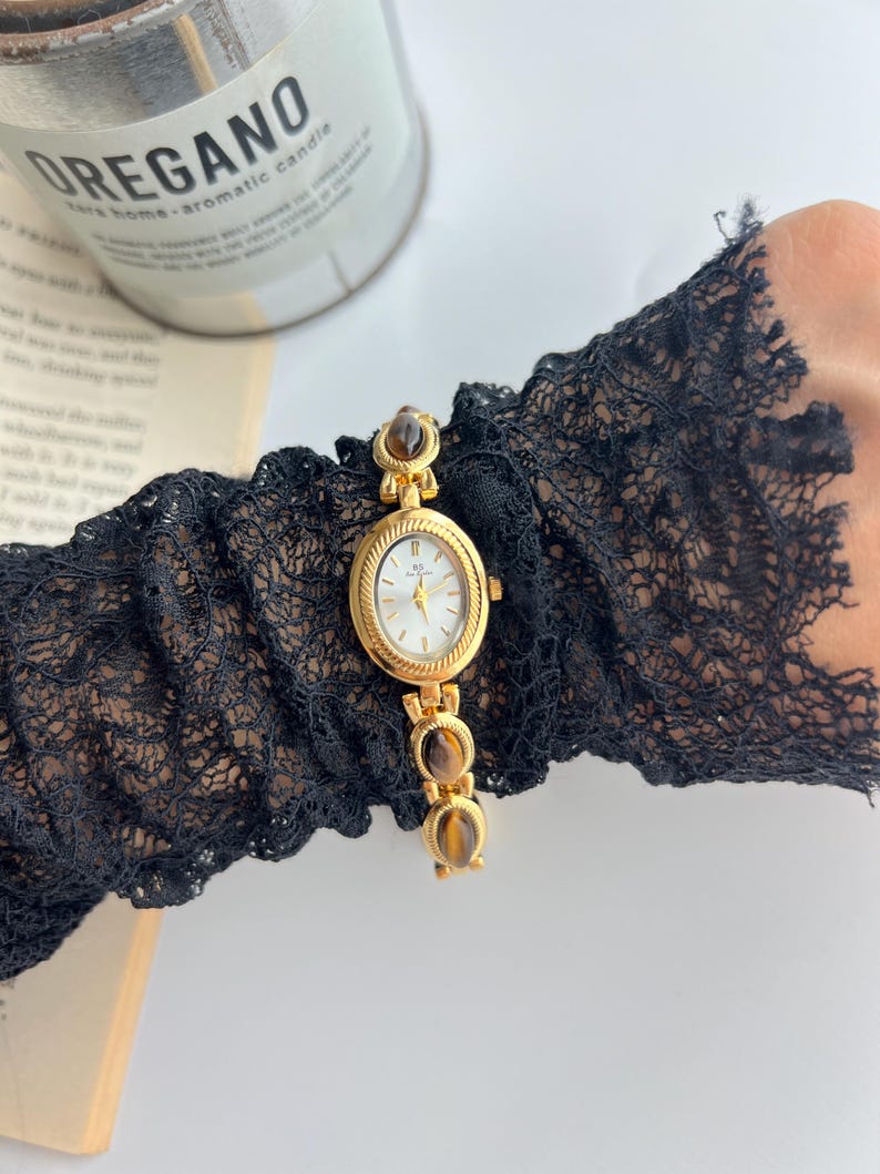 Puede incluir: Un reloj de pulsera dorado con esfera ovalada y esfera blanca. El reloj presenta piedras decorativas marrones y una correa dorada. El reloj se lleva en una manga de encaje negro. Una vela y un libro est&aacute;n en el fondo.