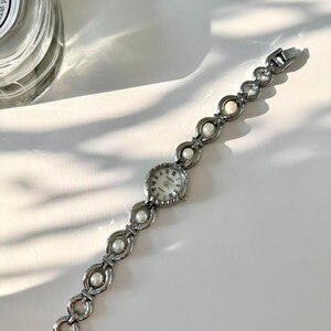 May include: A silver wristwatch with a round face and a linked bracelet-style strap. The strap features alternating circular links and faux pearls. The watch face has a white background with black numerals and hands. The watch is on a white surface.