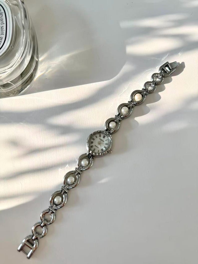 May include: A silver wristwatch with a round face and a linked bracelet-style strap. The strap features alternating circular links and faux pearls. The watch face has a white background with black numerals and hands. The watch is on a white surface.
