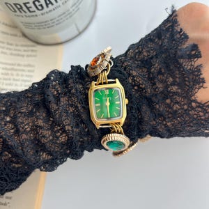 Puede incluir: Un reloj dorado con esfera verde y forma rectangular. El reloj presenta piedras preciosas decorativas de color naranja y verde. El reloj est&aacute; sujeto a una pulsera de cadena dorada. El reloj est&aacute; sobre una manga de encaje negro.