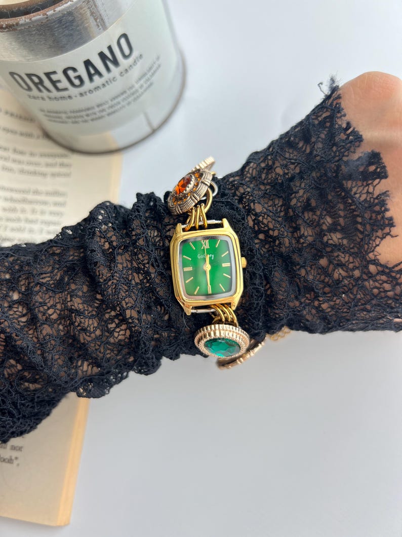 Puede incluir: Un reloj dorado con esfera verde y forma rectangular. El reloj presenta piedras preciosas decorativas de color naranja y verde. El reloj est&aacute; sujeto a una pulsera de cadena dorada. El reloj est&aacute; sobre una manga de encaje negro.