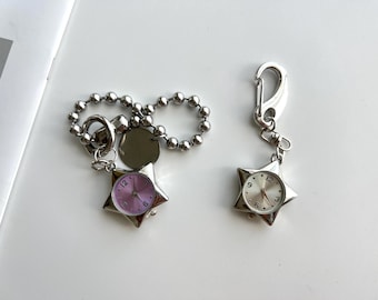 Könnte beinhalten: Zwei silberfarbene sternförmige Taschenuhren. Eine Uhr hat ein rosa Zifferblatt, die andere ein silbernes. Beide Uhren haben eine silberne Lünette und Zeiger. Eine Uhr ist an einer Kugelkette befestigt, die andere an einem Karabiner.