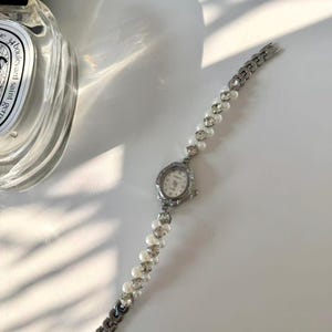 May include: A silver wristwatch with a small, oval face and a pearl and rhinestone strap. The watch face has a white background with black numerals and hands. The strap is made of alternating pearls and rhinestones, with a silver chain clasp.