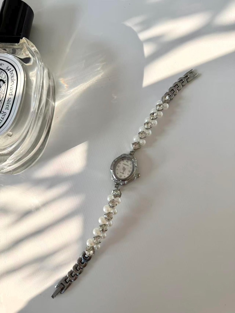 May include: A silver wristwatch with a small, oval face and a pearl and rhinestone strap. The watch face has a white background with black numerals and hands. The strap is made of alternating pearls and rhinestones, with a silver chain clasp.