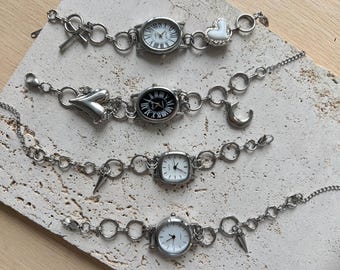 May include: Four silver-toned watch bracelets with different watch faces and decorative charms. One has a heart charm, another a crescent moon, and others have spike charms. The watch faces are round and square.