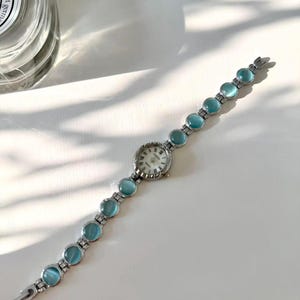 May include: A silver wristwatch with a round, white face and Roman numerals. The watch strap has light blue, circular stones. The watch is shown on a white surface. A classic design.