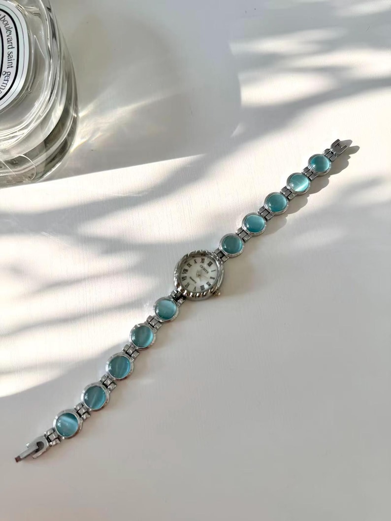 May include: A silver wristwatch with a round, white face and Roman numerals. The watch strap has light blue, circular stones. The watch is shown on a white surface. A classic design.